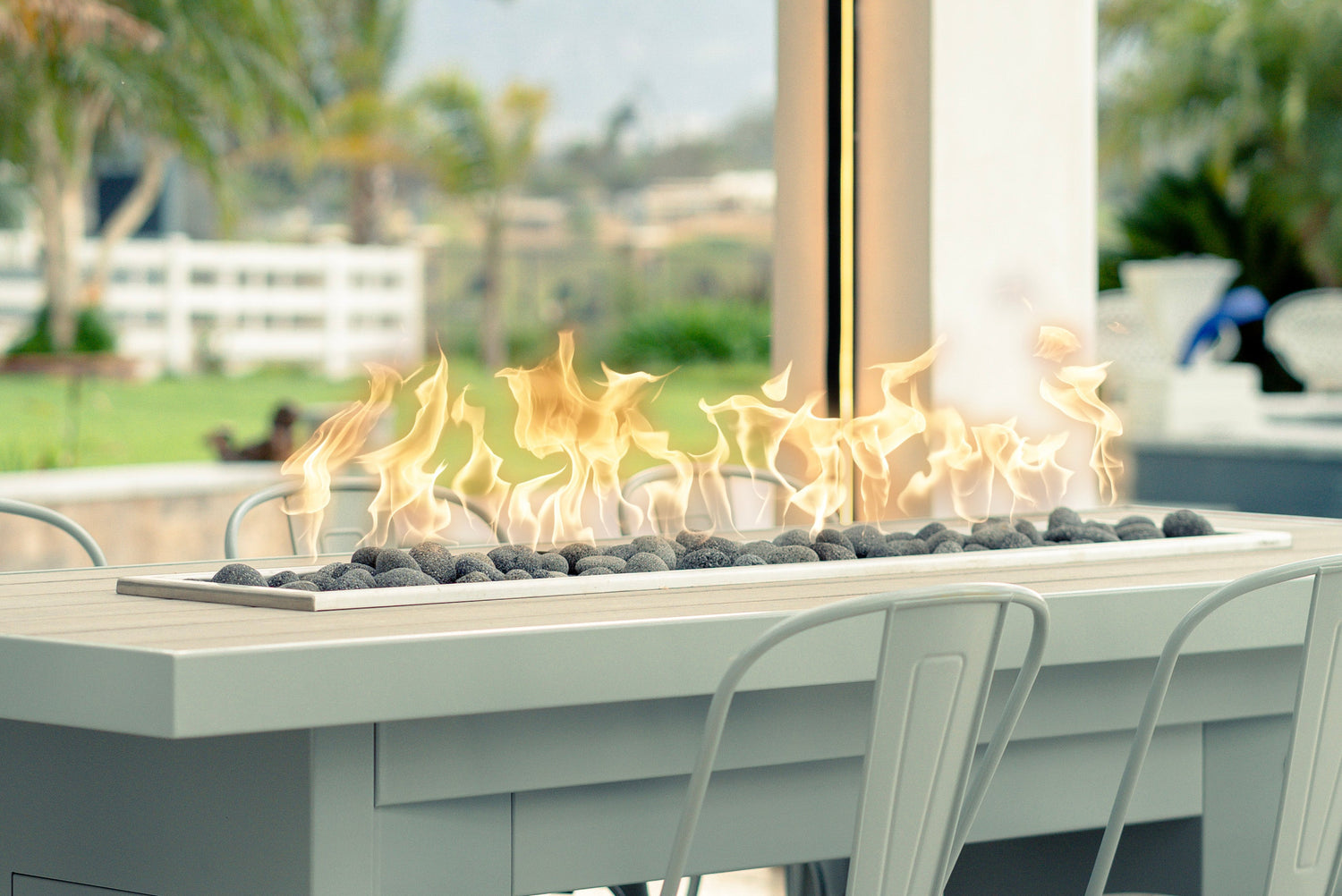 Modern rectangular fire pit table with flames over black fire stones, set on a patio dining area with outdoor chairs